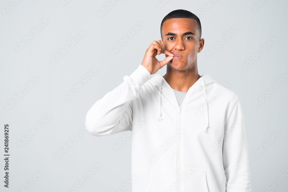 Dark-skinned young man with white sweatshirt showing a sign of closing mouth and silence gesture doing like closing his mouth with a zipper on grey background Dark-skinned young man with white sweatshirt showing a sign of closing mouth and silence gesture doing like closing his mouth with a zipper on grey background