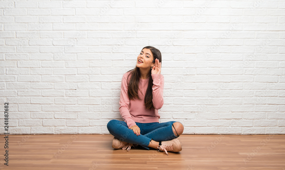 Teenager girl sitting on the floor in a room listening to something by putting hand on the ear