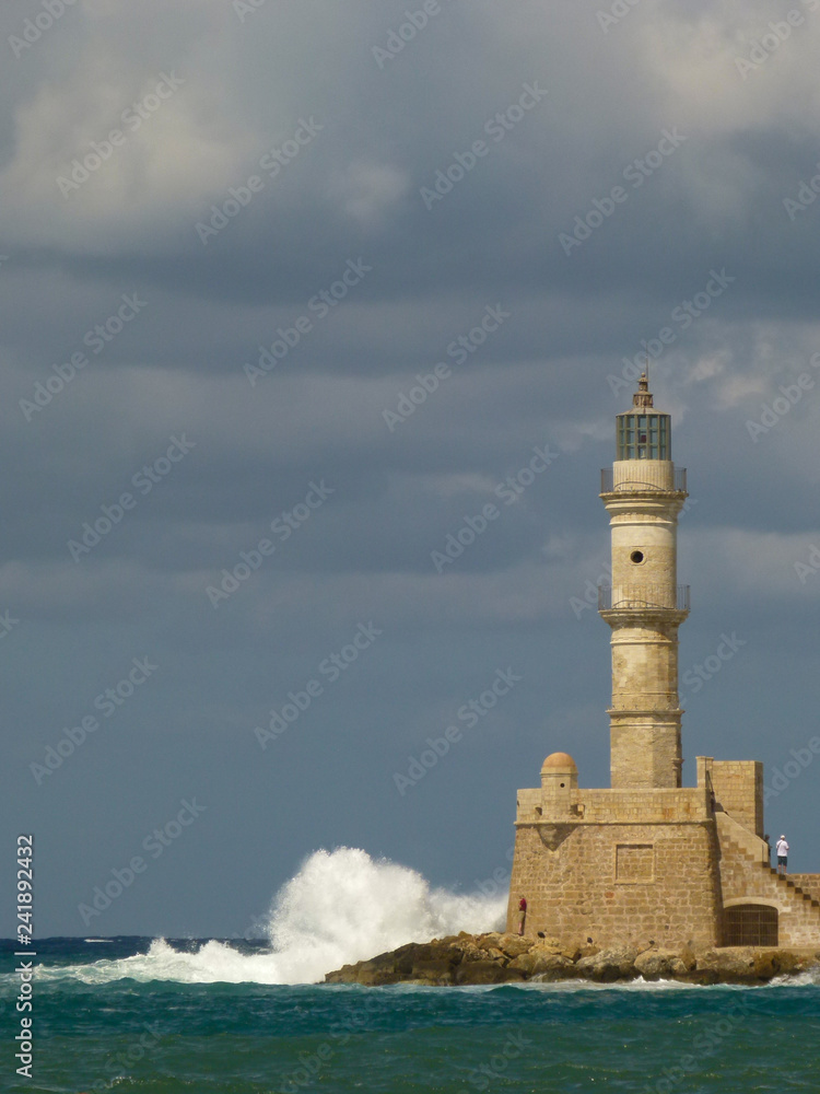 White waves with a lot of splashes crash into Chania lighthouse under a gray sky with clouds