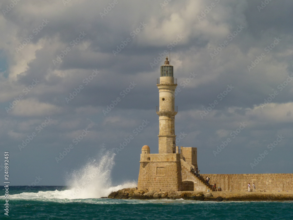 Gloomy gray clouds hang over the lighthouse of yellow and white bricks in Chania, about which waves crash
