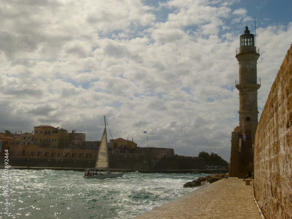 The yacht under the snow-white sail goes to the exit from the harbor of Chania at the lighthouse