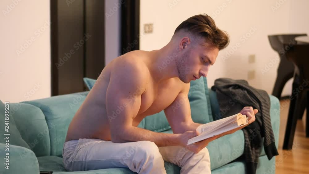 Handsome shirtless athletic young man reading book while sitting on couch