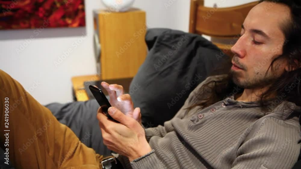 Man on his cellphone in bed. Man with long hair typing on his cellphone, lying in his bed in bedroom with painting - panning right Man on his cellphone in bed. Man with long hair typing on his cellphone, lying in his bed in bedroom with painting - panning right