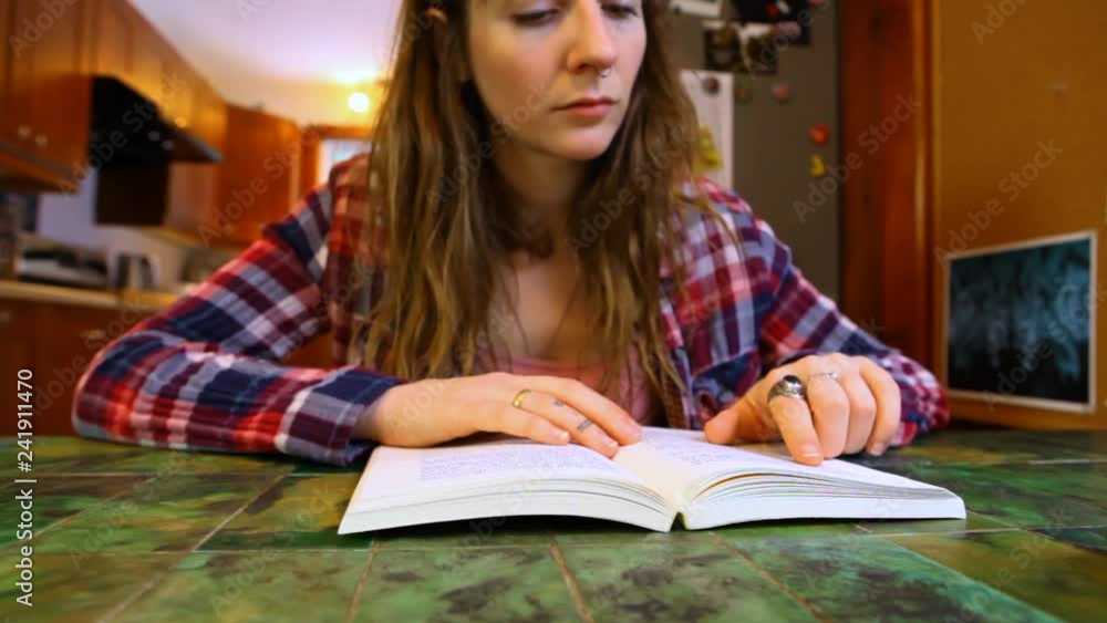 Woman is reading a book at the table. Young woman wearing checkered shirt is reading a book at her dining table. Fixed low angle from the table Woman is reading a book at the table. Young woman wearing checkered shirt is reading a book at her dining table. Fixed low angle from the table