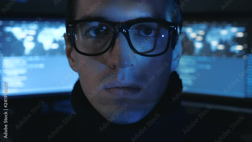 Portrait of Hacker programmer in glasses looks at camera while blue code characters reflect on his face in cyber security center filled with display screens. Portrait of Hacker programmer in glasses looks at camera while blue code characters reflect on his face in cyber security center filled with display screens.