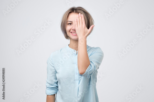 European young woman in blue shirt closing one eye with hand and smiling.