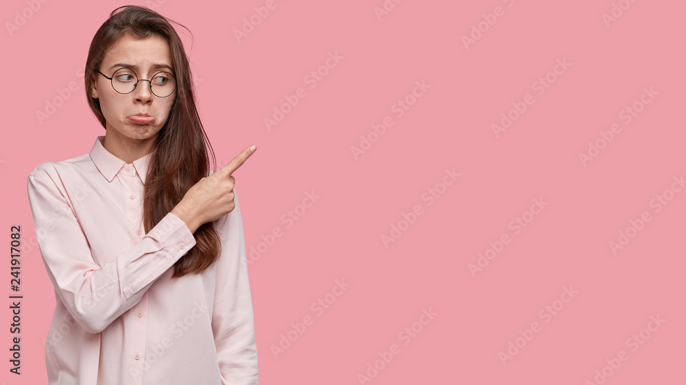 Unimpressed dejected woman purses lips, points aside upwards, being dejected, wears transparent glasses and shirt, stands over pink studio wall with blank space for your advertising content. Unimpressed dejected woman purses lips, points aside upwards, being dejected, wears transparent glasses and shirt, stands over pink studio wall with blank space for your advertising content.