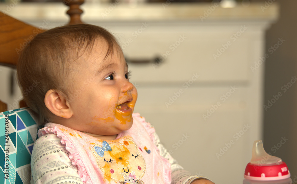 Fototapeta premium Infant laughing while eating dinner with food smeared on her face and down her bib