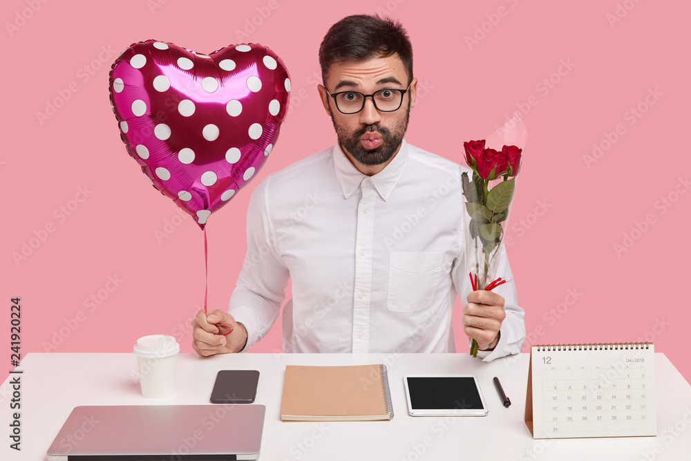Φωτογραφία Indoor shot of handsome unshaven man pouts lips, carries valentine and bouquet, has romantic relationships in office, poses over pink studio wall, busy working, uses modern gadgets