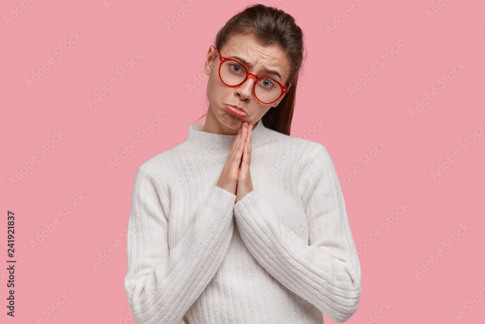 Portrait of pleading woman purses lips, keeps palms in praying gesture, asks for fortune, wears red spectacles, white casual jumper, models against pink background, has sad facial expression