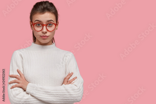 Studio shot of pretty woman looks with attentive facial expression, keeps hands crossed, wears jumper, stands over pink studio wall, blank space for your advertisement or promotional content photo