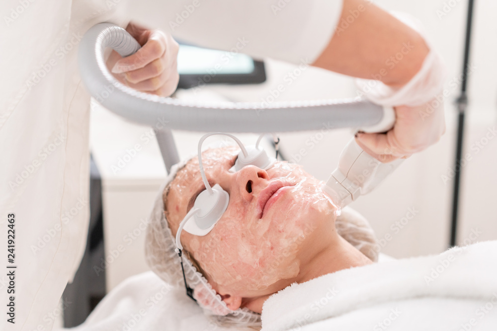 Naklejka premium Anti acne phototherapy with professional equipment. Beautiful woman in beauty salon during photo rejuvenation procedure. Laser face skin treatment at cosmetic clinic. Hardware cosmetology
