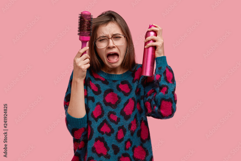 Displeased gloomy young woman cries from despair, makes hairdo, being in hurry, expresses negative emotions, wears loose sweater, uses hairspray, stands in beauty salon against pink background Displeased gloomy young woman cries from despair, makes hairdo, being in hurry, expresses negative emotions, wears loose sweater, uses hairspray, stands in beauty salon against pink background