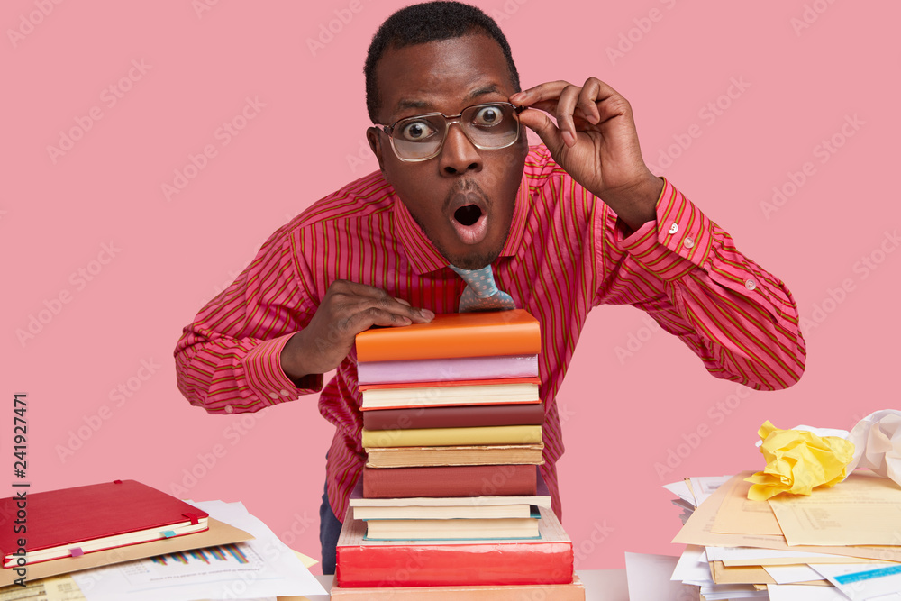 Isolated shot of amazed young black man looks scrupulously through spectacles, leans on pile of books, keeps hand on rim of glasses, dressed formally, isolated over pink background. Omg concept Isolated shot of amazed young black man looks scrupulously through spectacles, leans on pile of books, keeps hand on rim of glasses, dressed formally, isolated over pink background. Omg concept