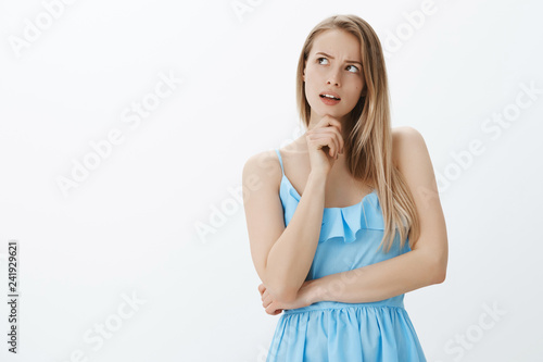 Concerned unsure and heistant young attractive woman with fair hair in blue dress open mouth touching chin and looking at upper left corner as thinking  being uncertain and confused how act right