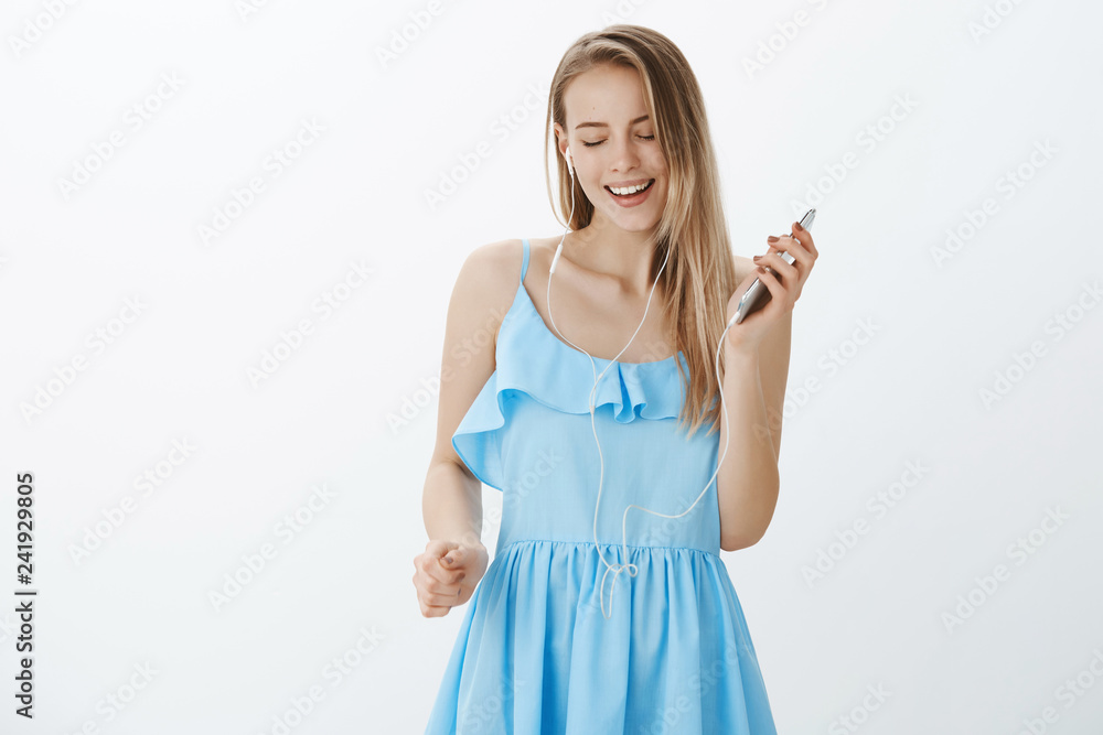 Girl forgetting all troubles as enjoying cool music listening in app via smartphone, wearing earphones singing along and smiling broadly, liking beats in earbuds feeling awesome over gray background Girl forgetting all troubles as enjoying cool music listening in app via smartphone, wearing earphones singing along and smiling broadly, liking beats in earbuds feeling awesome over gray background