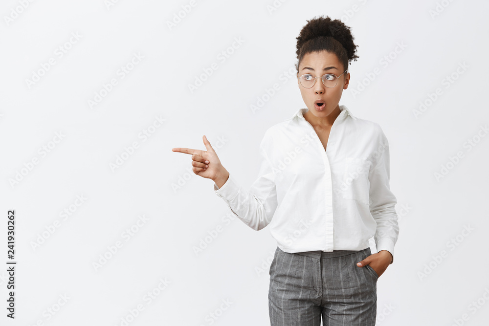Studio shot of impressed and surprised attractive dark-skinned female with curly hair in glasses and white collar shirt, holding hand in pocket, folding lips from curiosity, looking and pointing left Studio shot of impressed and surprised attractive dark-skinned female with curly hair in glasses and white collar shirt, holding hand in pocket, folding lips from curiosity, looking and pointing left