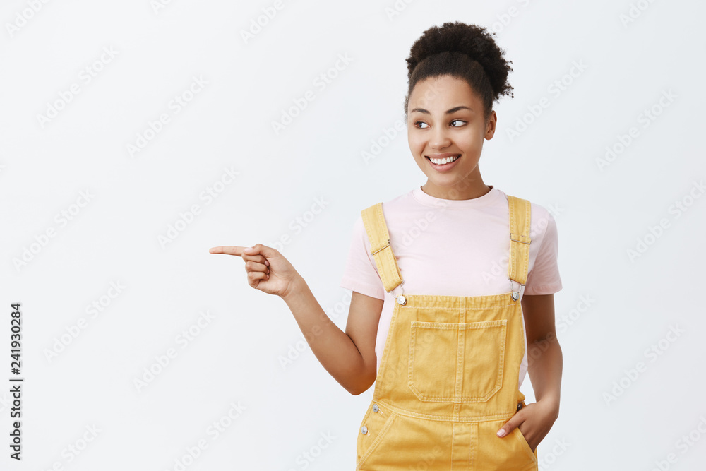 Cute young and tender female sibling with dark skin in yellow trendy overalls, holding hand in pocket, pointing and looking left while telling direction to friend who trying find bathroom Cute young and tender female sibling with dark skin in yellow trendy overalls, holding hand in pocket, pointing and looking left while telling direction to friend who trying find bathroom