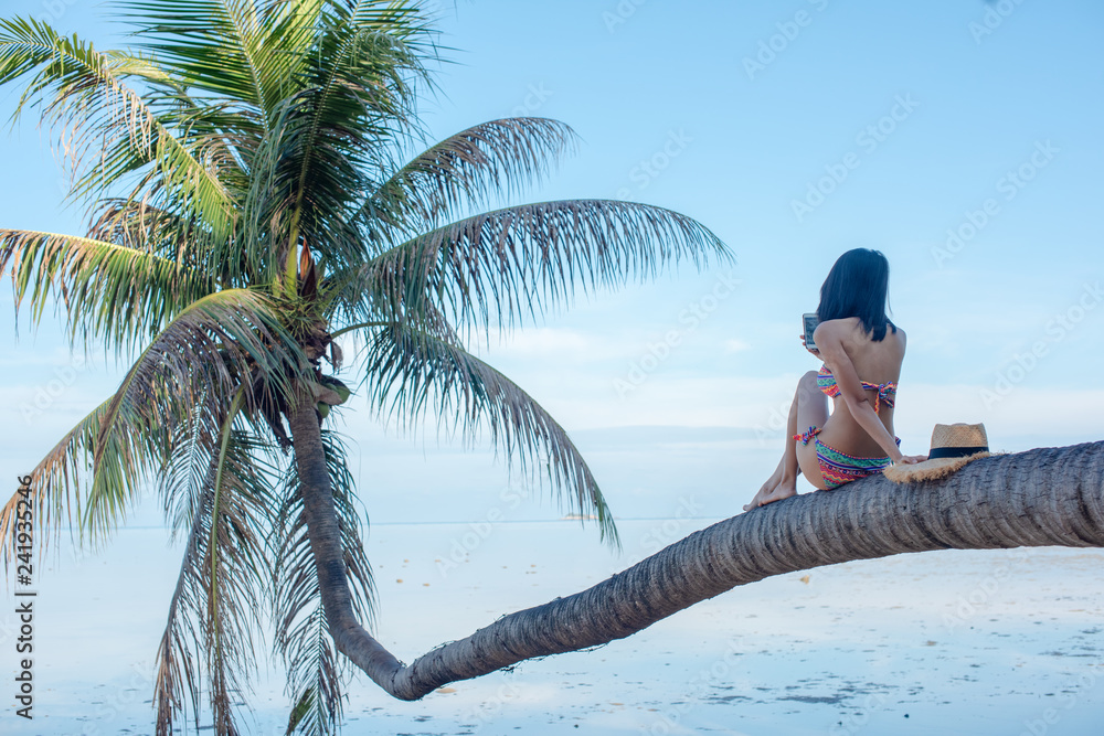 Fototapeta premium woman in bikini enjoy the seabeach by sitting onto the laying of coconut plam tree over the sea water in daylight