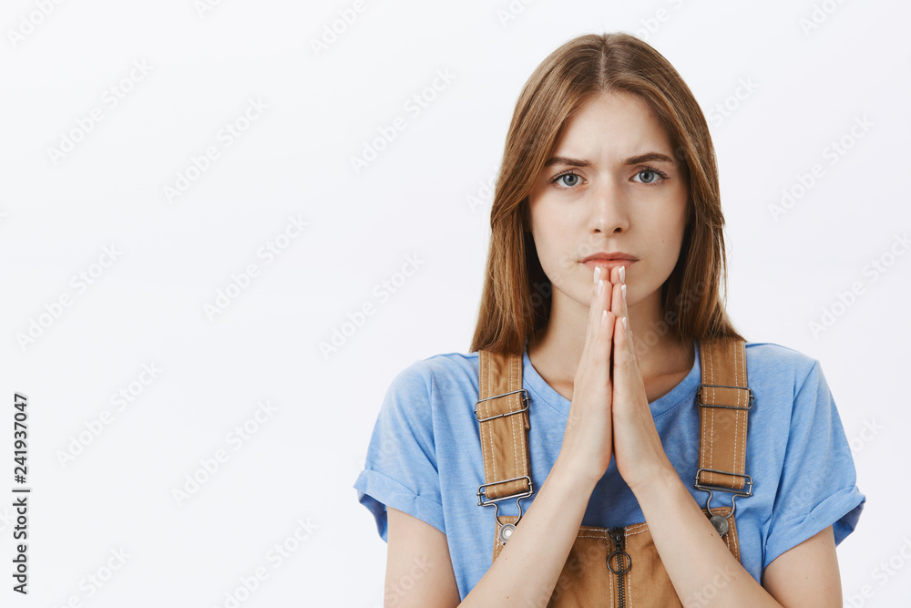 Close-up shot of hopeful serious-looking determined cute young woman with chestnut hair standing concerned holding hands in pray looking focused at camera with faith and hope over gray wall Close-up shot of hopeful serious-looking determined cute young woman with chestnut hair standing concerned holding hands in pray looking focused at camera with faith and hope over gray wall