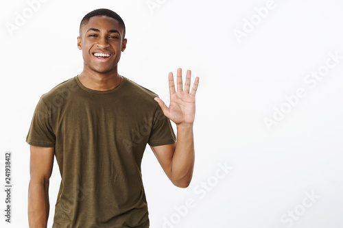 Friendly attractive nice african american male friend raising palm to wave smiling broadly saying hello, greeting friends in cafe welcoming or inviting people to party, posing over gray background