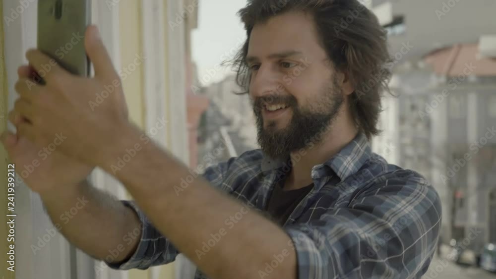 Smiling bearded man standing on balcony and looking at smartphone.