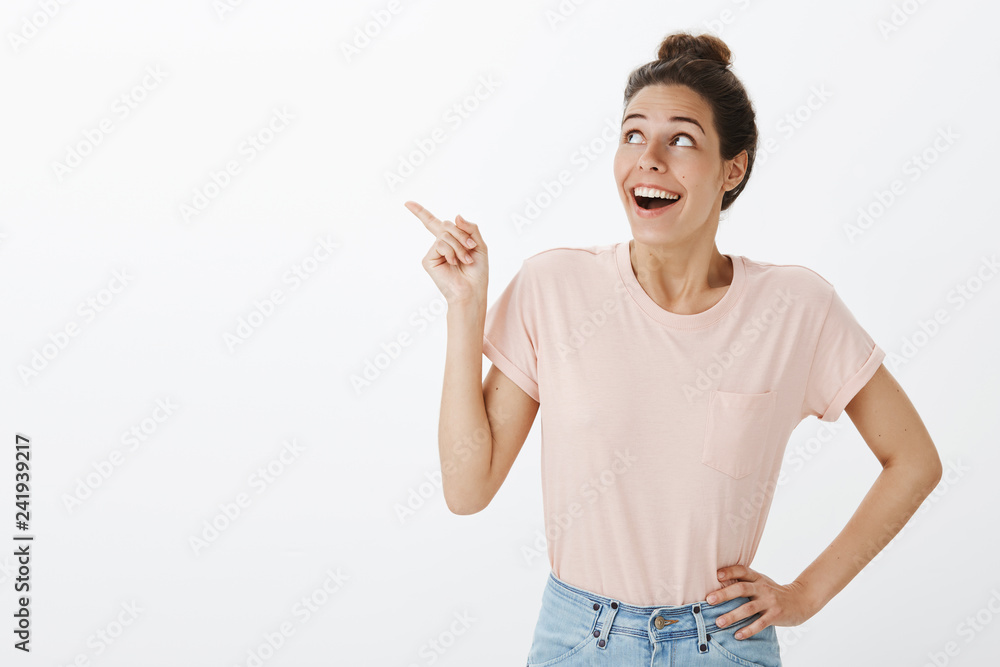 Enthusiastic impressed and happy young joyful woman in t-shirt and jeans dropping jaw from joy and amazement pointing, looking at upper left corner excited and surprised over grey wall Enthusiastic impressed and happy young joyful woman in t-shirt and jeans dropping jaw from joy and amazement pointing, looking at upper left corner excited and surprised over grey wall