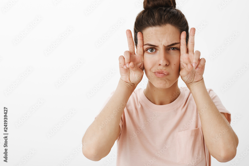 Fototapeta premium Girl making faces as feeling gloomy trying cheer up fooling around frowning and smirking showing peace gesture sideways standing serious and hilarious over gray background in trendy t-shirt