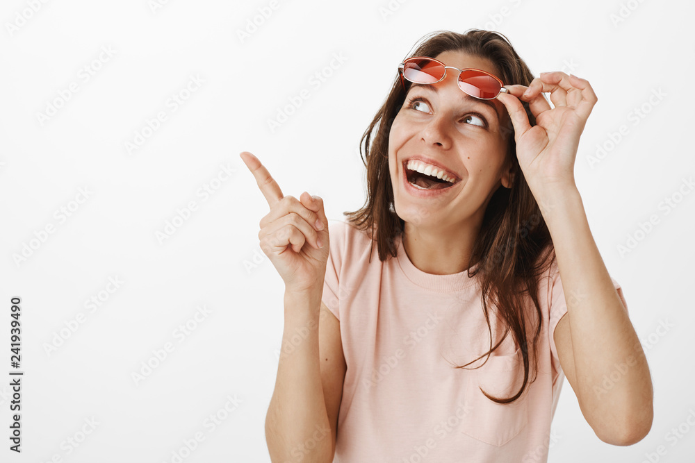 Amused girl looking at funny cat on tree loving animals taking off  sunglasses as observing pointing at upper left corner smiling and laughing  amazed and joyful, having fun against gray background Stock