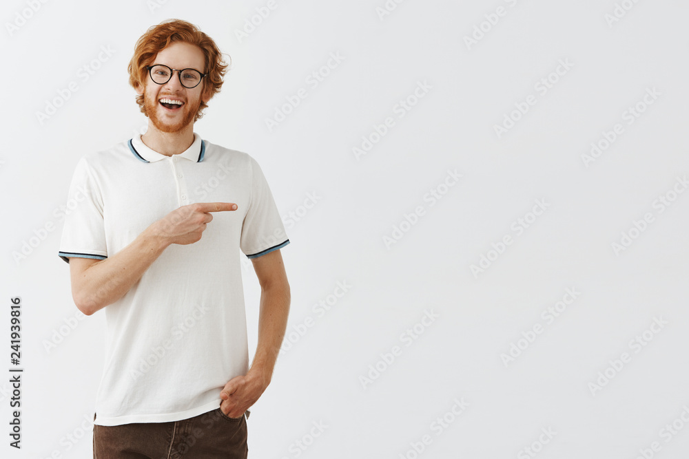 Happy enthusiastic handsome redhead guy with long beard in glasses and casual outfit pointing right with forefinger and smiling broadly while being uplifted and suggesting friends stay in his place Happy enthusiastic handsome redhead guy with long beard in glasses and casual outfit pointing right with forefinger and smiling broadly while being uplifted and suggesting friends stay in his place