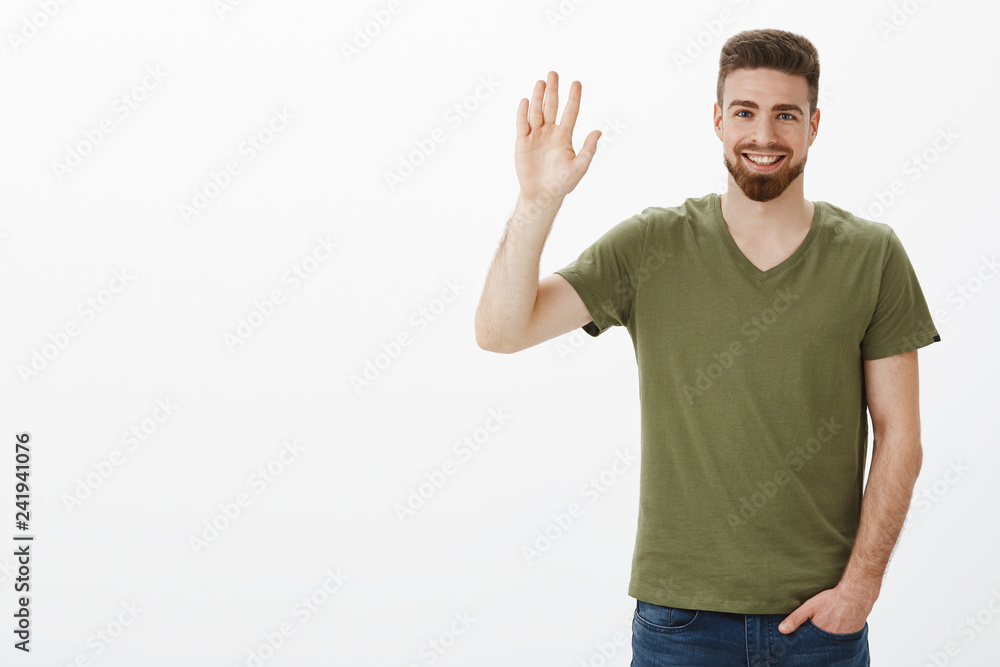 Friendly-looking charming bearded boyfriend feeling shy waving with raised hand in hello or hi gesture, holding hand in pocket casually as meeting new people, greeting over white background
