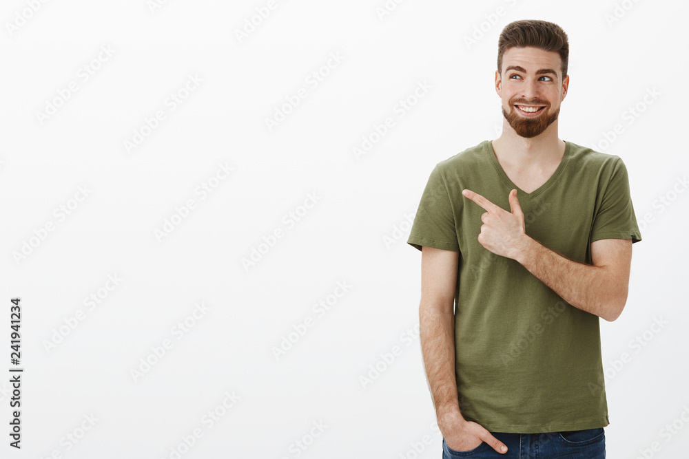 Indoor shot of smart handsome bearded caucasian guy in good mood happy smiling delighted as looking and pointing at upper left corner having great plan pleased against white background Indoor shot of smart handsome bearded caucasian guy in good mood happy smiling delighted as looking and pointing at upper left corner having great plan pleased against white background