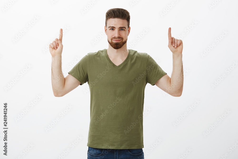 Naklejka premium Guy assured you need check out product, pointing with raised index fingers up at copy space with confident smirk, looking strict and self-assured at camera encouraging try it over white background