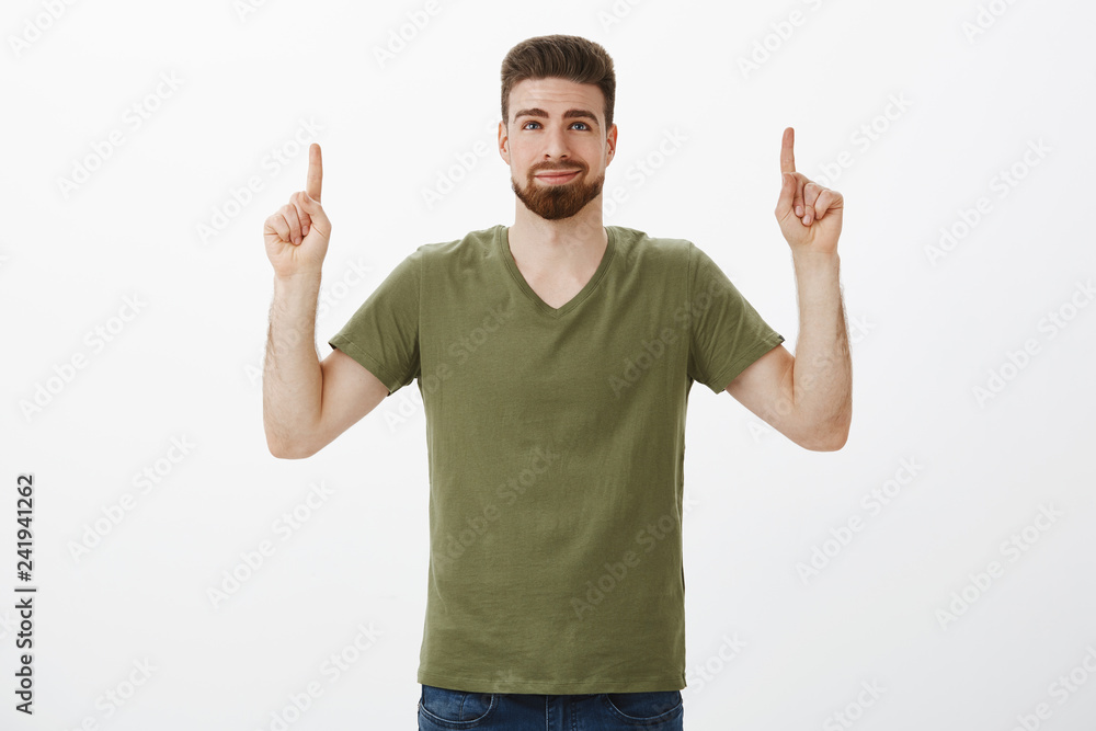 Fototapeta premium Portrait of dreamy delighted and cute bearded boyfriend in love smirking pleased and releaved looking, pointing up with raised hands as finding awesome gift for valentines day against white background