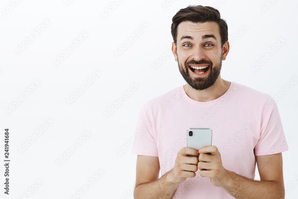 Waist-up shot of excited happy and enthusiastic handsome adult man playing games in smartphone holding cellphone gazing satisfied and joyful at camera with broad smile posing against gray wall Waist-up shot of excited happy and enthusiastic handsome adult man playing games in smartphone holding cellphone gazing satisfied and joyful at camera with broad smile posing against gray wall