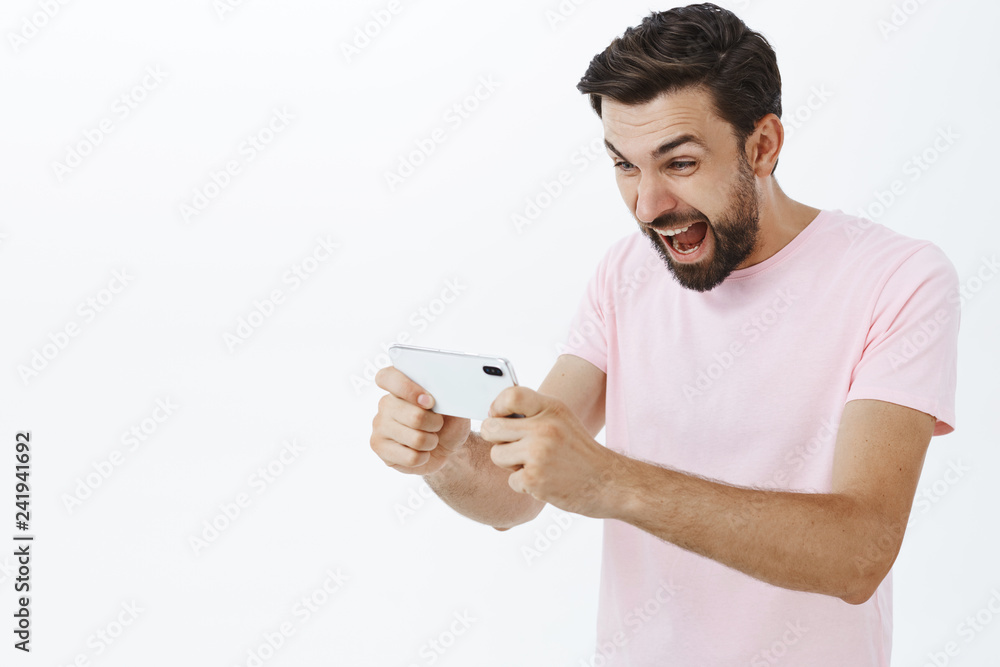 Waist-up shot of excited and thrilled male gambler with beard and cool hairstyle yelling cheerful and joyful holding smartphone with passion playing game posing against gray background delighted Waist-up shot of excited and thrilled male gambler with beard and cool hairstyle yelling cheerful and joyful holding smartphone with passion playing game posing against gray background delighted