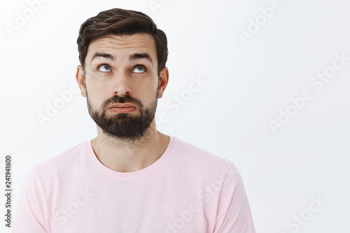 Close-up shot of unlucky, tired and upset attractive young bearded male with beard raising eyebrows in despair looking up with regret and disappointment, standing unconfident and exhausted