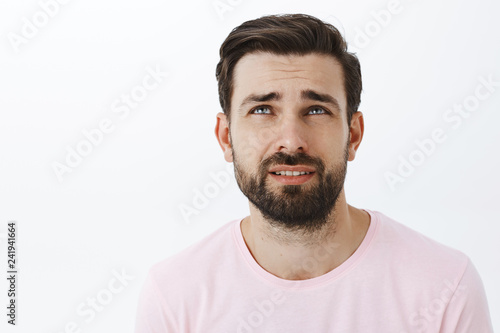 Waist-up shot of gloomy and unhappy adult bearded man with blue eyes frowning looking up bothered with regret standign displeased and concerned over gray wall overthinking feeling sad