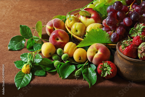 Ripe peaches with leaves and flowers on the table with a brown tablecloth.
