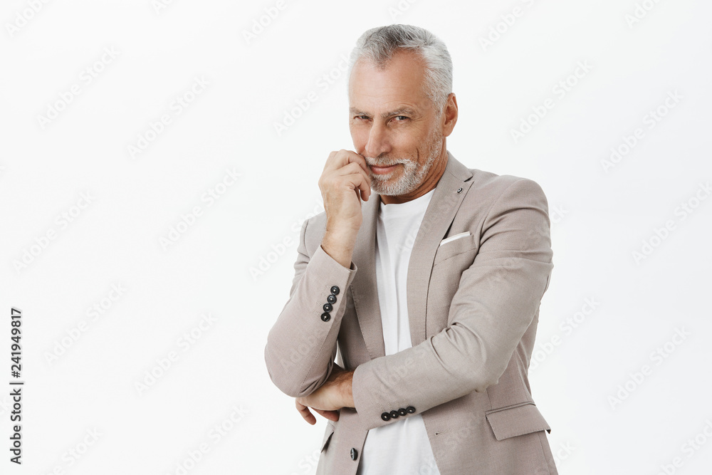 Smart and tricky enthusiastic old man with white beard and hair crossing hand against chest and touching lip smiling curiously and squinting from interest at camera having idea or intention Smart and tricky enthusiastic old man with white beard and hair crossing hand against chest and touching lip smiling curiously and squinting from interest at camera having idea or intention