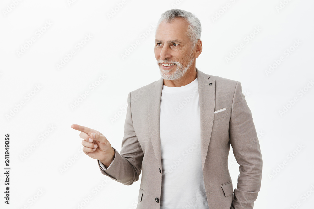 Realxed and amused good-looking senior male entrepreneur in grey suit over t-shirt looking and pointing left with happy satisfied smile liking new product of company presented on copy space Realxed and amused good-looking senior male entrepreneur in grey suit over t-shirt looking and pointing left with happy satisfied smile liking new product of company presented on copy space