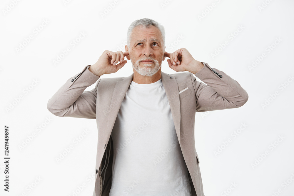 Portrait of old businessman being disturbed by loud noise comming upwards closing ears with index fingers like earplugs looking up worried and displeased wish sound or yell over white background Portrait of old businessman being disturbed by loud noise comming upwards closing ears with index fingers like earplugs looking up worried and displeased wish sound or yell over white background