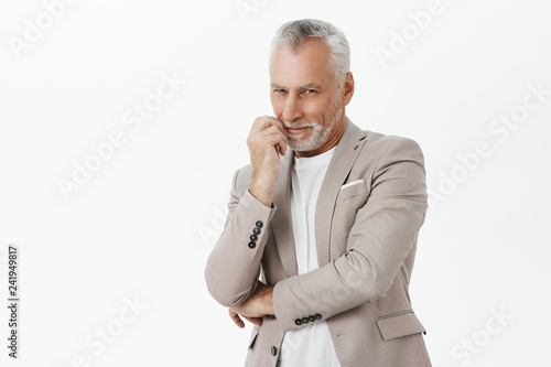 Smart and tricky enthusiastic old man with white beard and hair crossing hand against chest and touching lip smiling curiously and squinting from interest at camera having idea or intention