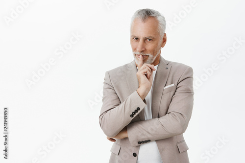 Enthusiastic and creative charming senior businessman with white hair and moustache in elegant stylish grey suit showing shh gesture with index finger over mouth smiling tricky having plan or surprise