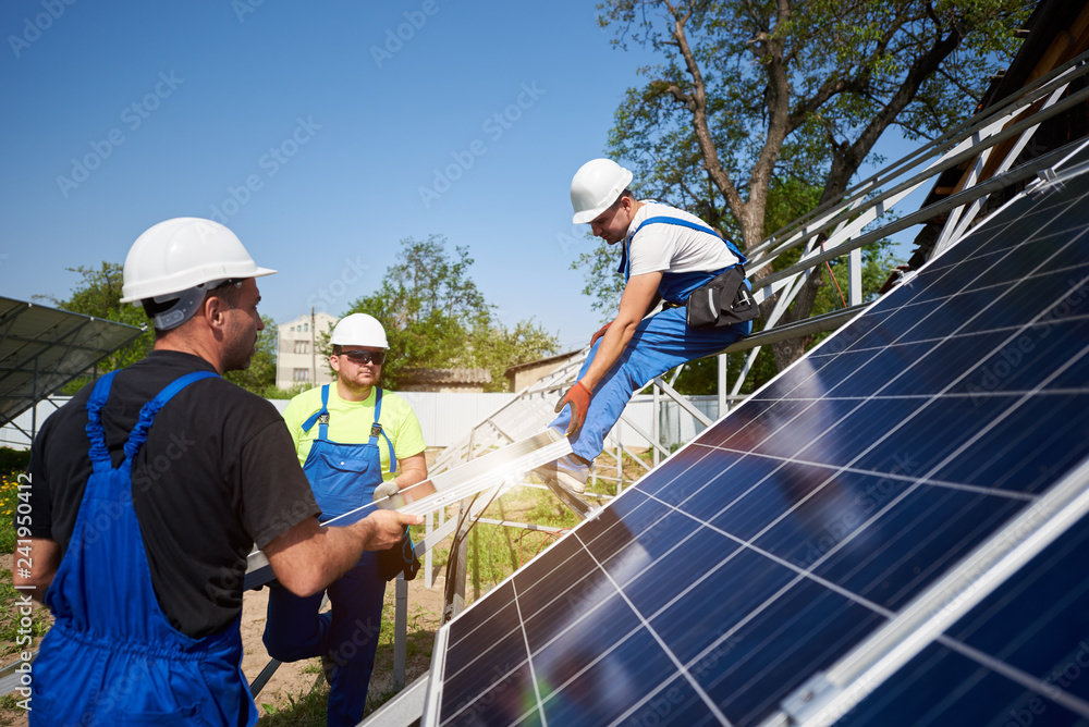 Team of three professional workers lifting heavy solar photo voltaic panels on high steel platform. Exterior solar system installation, alternative renewable green energy generation concept.