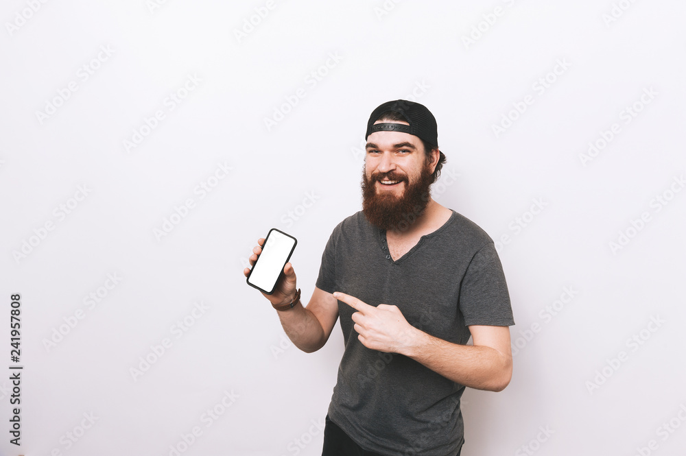 Photo of cheerful bearded man in hat and shrt, poiting to white screen of smartphone Photo of cheerful bearded man in hat and shrt, poiting to white screen of smartphone