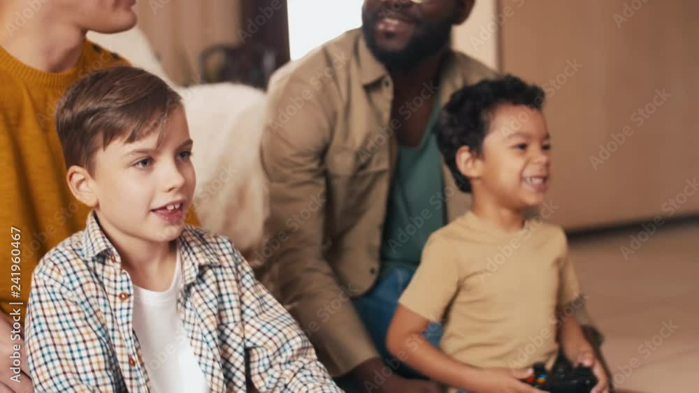 Two multi-ethnic friends discussing plans for weekend while their sons sitting near and using joysticks to play video games Two multi-ethnic friends discussing plans for weekend while their sons sitting near and using joysticks to play video games