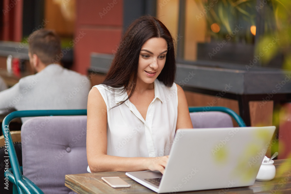 Pleased brunette woman in white clothes sits in front of opened laptop, reads response, recieves email on gadget, works as journalist, poses at desk in outdoor cafeteria connected to wireless internet Pleased brunette woman in white clothes sits in front of opened laptop, reads response, recieves email on gadget, works as journalist, poses at desk in outdoor cafeteria connected to wireless internet