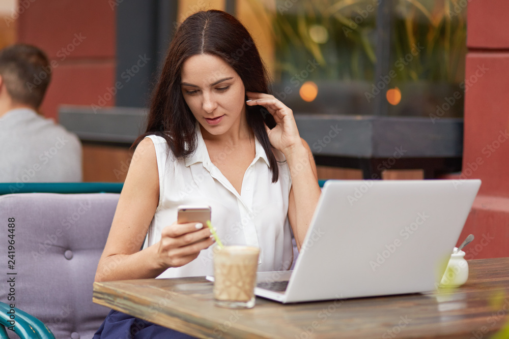 Horizontal shot of busy female freelancer holds mobile phone, text messages online, uses laptop computer, drinks milkshake, poses in outdoor restaurant, dressed in white clothes, sits in front of pc Horizontal shot of busy female freelancer holds mobile phone, text messages online, uses laptop computer, drinks milkshake, poses in outdoor restaurant, dressed in white clothes, sits in front of pc