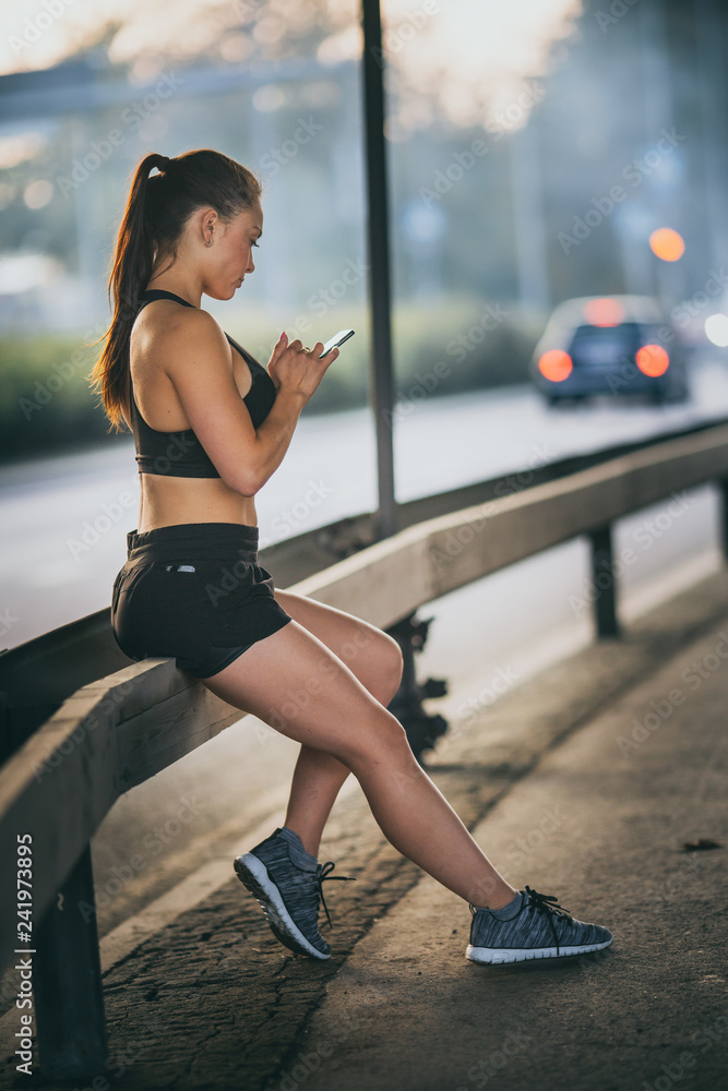 Shot of a Beautiful Confident Fitness Girl in Black Athletic Top is Using a Smartphone on a Street. She is in an Urban Environment Under a Bridge.